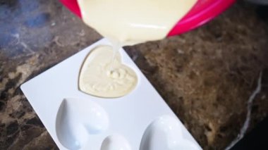 Kırmızı kaseden beyaz kremalı beyaz çikolatayı mutfak tezgahına beyaz kalp şeklinde bir kalıba döküyordu. Yüksek kalite 4k görüntü