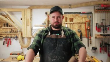 Elinde koruyucu bir önlükle beyaz, genç bir marangoz bir çekiç ve elleri çaprazlanmış bir keski tutuyor ve birçok aletle gülümsüyor. Yüksek kalite 4k görüntü