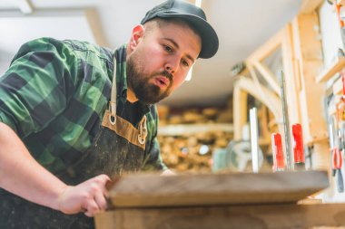 Genç erkek marangoz bir tahta parçasına talaş üfleyerek ahşabı temizliyor. Tahta ürünleri. Yüksek kalite fotoğraf
