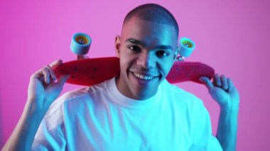 Beyaz tişörtlü mutlu Afro-Amerikalı çocuk sırtında bir kaykay tutuyor, pembe arka plan resmi. Yüksek kalite fotoğraf