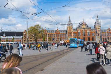 Amsterdam Centraal İstasyonu güneşli bir günde çekildi. Mavi tramvay ortaya gidiyor. Açık ceketli turist grupları geçiyor. Yüksek kalite fotoğraf