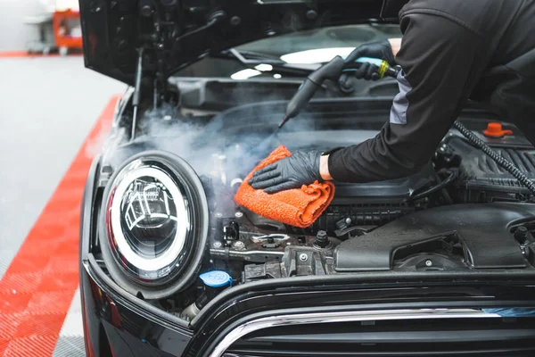 Araba temizleme konsepti. Buharla elle motor temizliği. Araba temizleme uzmanı siyah aracı temizliyor ve araba yüzeyini turuncu mikrofiber bezle siliyor. Yüksek kalite fotoğraf