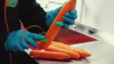 Worker in black uniform wearing protective gloves processing fresh carrots using vegetable peeler with kitchen sink in background. Preparing fresh products concept. High quality 4k footage