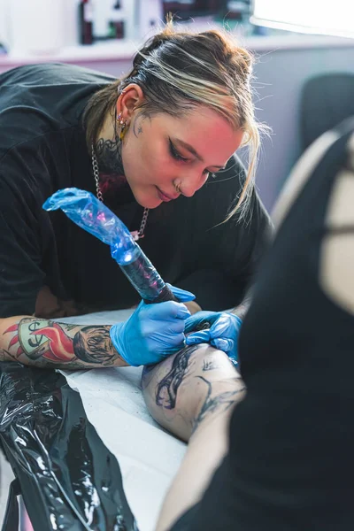 在她的作品中 女性纹身艺术家的垂直镜头 在纹身工作室忙着呢拿着纹身枪在腿上画纹身的女孩高质量的照片 — 图库照片