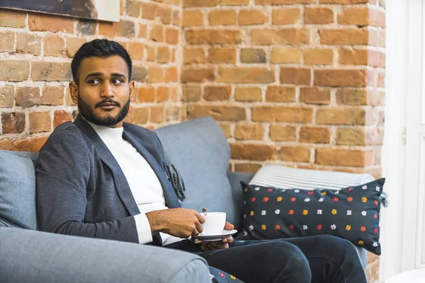 Indian man in a suit sitting on a sofa at a salon holding a cup of coffee against a brick wall. Looking away. High quality photo