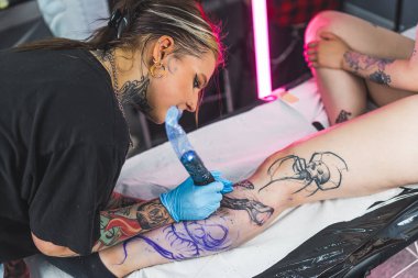 Dövme makinesi tutan ve müşterinin bacağına dövme çizen bir kadın. Baldır dövmesi. Çalışırken odaklanmış kadın sanatçı. Yüksek kalite fotoğraf