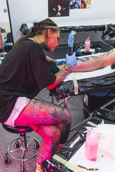 Tattooed artist focused on work - drawing a tattoo on her clients leg. Black ink tattoo. Vertical indoor shot. Tattooing studio. High quality photo