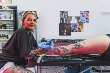 Mutlu kadın dövme sanatçısı müşterisinin bacağına dövme çizerken kameraya bakıyor ve gülümsüyor. Yüksek kalite fotoğraf