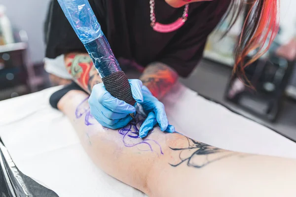Female tattooist holding professional tattooing gun and drawing tattoo on a female customers leg. Closeup shot. High quality photo