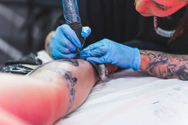 Focused tattoo artist in blue protective rubber gloves tattooing the leg of her client. Modern body art. Copy space. High quality photo