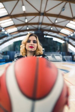 Beyaz bir kadın, kapalı bir sahada kameraya basketbol topunu gösteriyor. Yüksek kalite fotoğraf