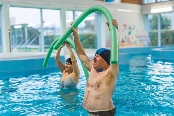 Indoor pool fitness exercises. Adult caucasian seniors doing aerobics exercises with foam pool noodles. High quality photo