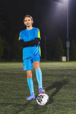 Mavi spor kıyafetli genç beyaz bir kız stadyumda futbol topuyla poz veriyor, tam dikey. Yüksek kalite fotoğraf