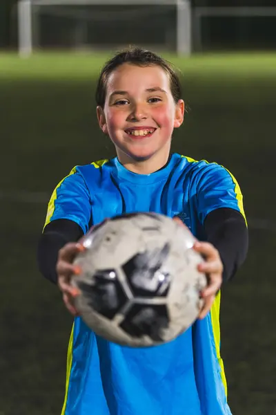 medium shot of a joyful teenage girl holding a soccer ball toward a camera, late evening practice. High quality photo