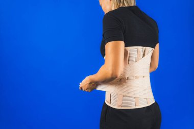 Sırtındaki korseyi sırtındaki acıdan ve omurgadan korumak için düzelten bir kadın. Mavi arka planda arka plan. Yüksek kalite fotoğraf