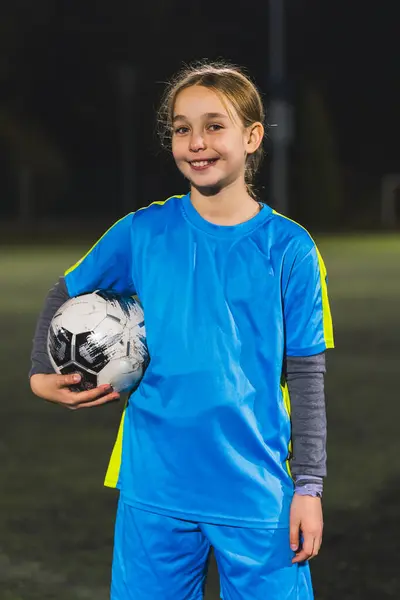 medium vertical shot of a soccer player little girl holding a ball in one hand and smiling at the camera. High quality photo