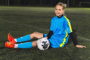 Futbolcu Kafkasyalı kız elinde topla sahada oturuyor ve gülümsüyor, geceleri pratik yapıyor. Yüksek kalite fotoğraf