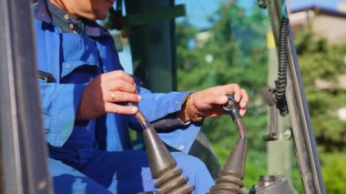 Yakından bakınca mavi üniformalı bir adamın traktör sürdüğünü görüyoruz. İşçi konsepti, inşaat alanında kazıcı kullanan erkek operatör. Yüksek kalite 4k görüntü