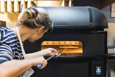 Önlüklü kadın pizzayı kabuklu elektrikli fırına koyuyor, restoranın mutfağında çalışıyor. Yüksek kalite fotoğraf