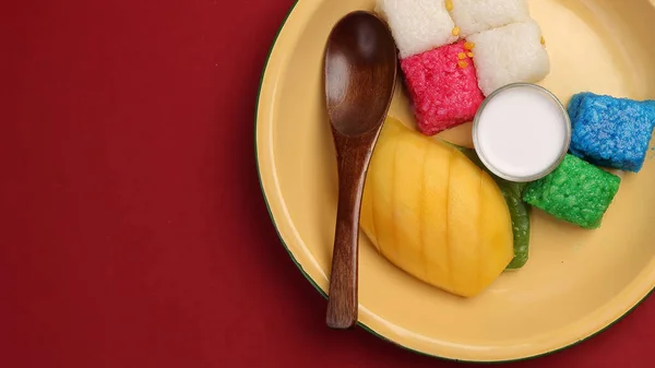 Thai sweet desert red blue yellow white mango sticky rice with coconut cream milk on enamel tin metal plate wood spoon red background