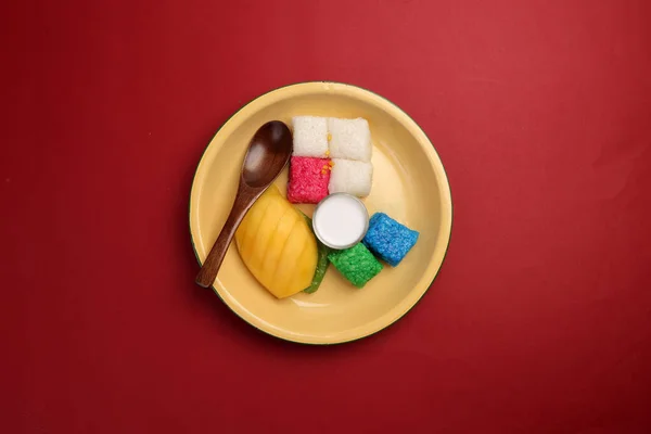 Thai sweet desert red blue yellow white mango sticky rice with coconut cream milk on enamel tin metal plate wood spoon red background