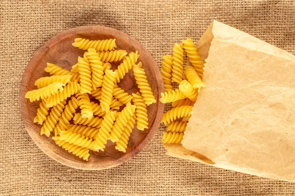 Durum wheat pasta on a wooden saucer and in a kraft paper bag on a jute cloth, macro, top view.