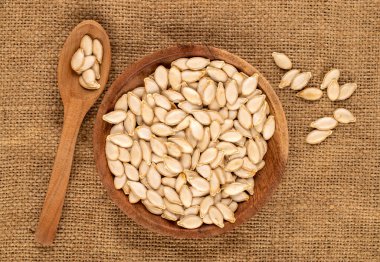 Unpeeled raw pumpkin seeds in wooden plate with spoon on jute cloth, macro, top view. clipart