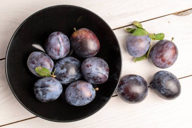 Birkaç olgun siyah erik siyah seramik tabakta, yakın planda, beyaz ahşap bir masada, üst tarafta..
