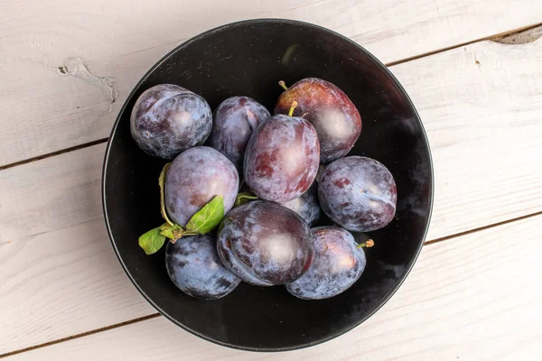 stock image Several ripe black plums in a black ceramic plate, close-up, on a white wooden table, top view.