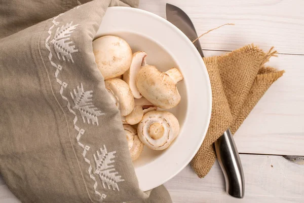 Several organic fresh appetizing mushrooms champignon in a white ceramic plate, with a metal knife and two linen and jute napkins, on a table made of natural wood, top view.