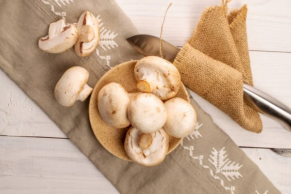 A few whole and two halves of organic fresh appetizing mushrooms champignon with a ceramic saucer, with a metal knife and two linen and jute napkins, on a table made of  natural wood, top view.