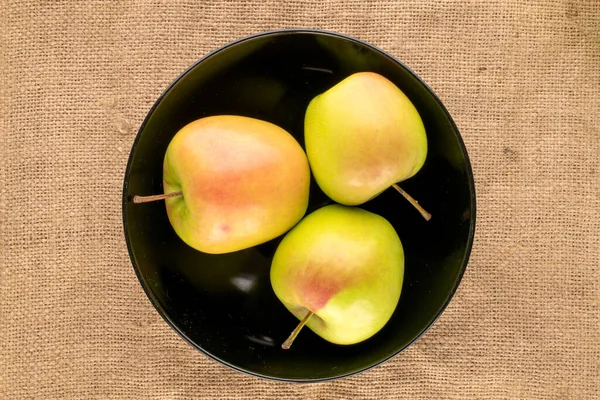 Three sweet green apples in a black plate on a jute cloth, macro, top view.