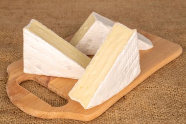 Three slices of delicious brie cheese with wooden kitchen board on jute cloth, macro.