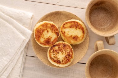Two homemade cheese pancakes on ceramic saucer with white napkin and two cups on wooden table, macro, top view.