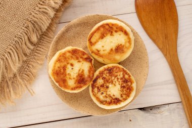 Three homemade cheese pancakes on ceramic saucer with jute napkin, wooden spoon on wooden table, macro, top view.