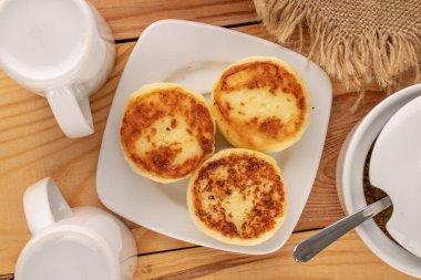 Three homemade cheese pancakes on ceramic saucer with jute napkin, white sugar bowl and two cups on wooden table, macro, top view.