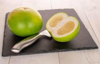 One whole and half organic Citrus Sweetie on a slate board with a metal knife, close-up, on a white wooden table.