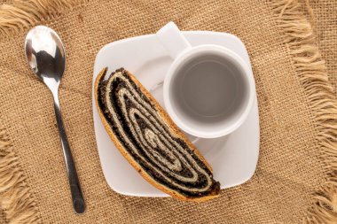 One piece of appetizing poppy seed roll on white ceramic plate with cup and metal spoon on jute cloth, macro, top view.