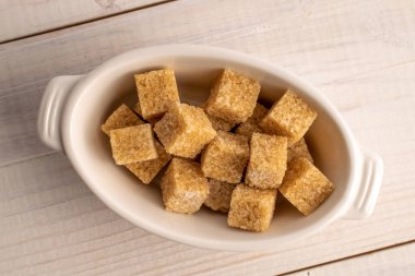 Several brown sugar cubes in a white ceramic bowl, close-up, on a white wooden table.