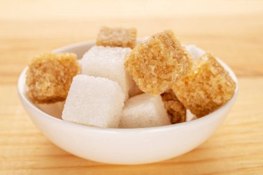 Several cubes of brown and white sugar in a white ceramic bowl, close-up, on a wooden table.