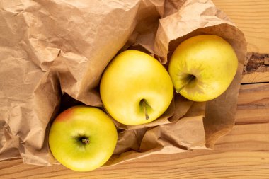 Three yellow organic apples in a paper bag on a wooden table, macro, top view.