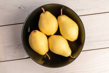 Four ripe yellow pears in a black ceramic plate, close-up, on a white wooden table, top view.