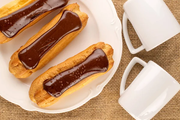 stock image Three chocolate eclairs with a white ceramic plate and two cups on a jute cloth, close-up, top view.