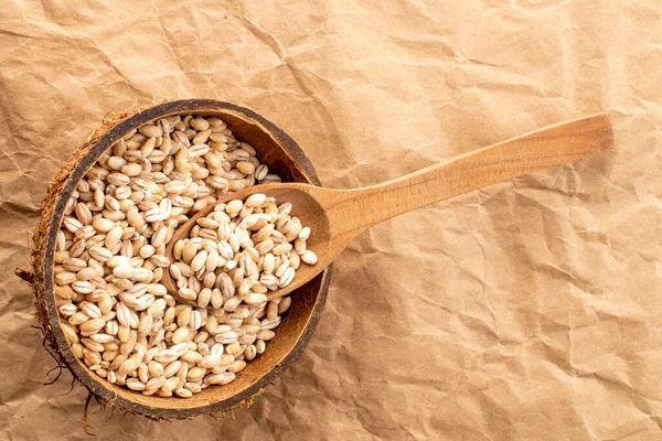 Organic pearl barley in coconut shell with wooden spoon on kraft paper, close-up, top view.