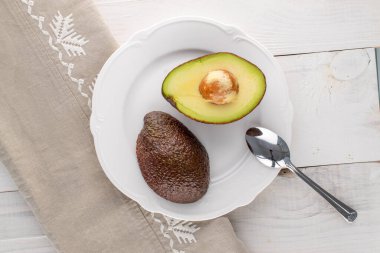 Seramik bir tabakta iki yarım organik avokado, ahşap bir masada metal kaşık, yakın çekim, üst manzara..