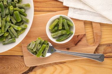 Beyaz seramik tabakta donmuş yeşil fasulyeler, tahta bir masada metal kaşık, makro, üst manzara..