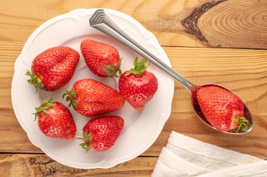 Beyaz seramik tabağı olan birkaç tatlı çilek, metal kaşık ve tahta masa üzerinde beyaz kağıt peçete, makro, üst manzara..