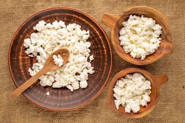 stock image Fresh cottage cheese in a ceramic plate, two wooden cups and a wooden spoon on a jute cloth, macro, top view.