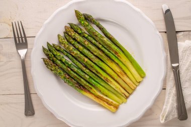 Seramik beyaz bir tabakta, ahşap bir masada, çatal ve bıçakla ızgara kuşkonmaz, makro, üst manzara..