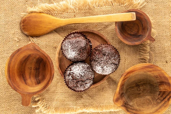 Three homemade chocolate cupcakes with wooden saucer, three cups, wooden spoon and jute napkin on jute cloth, macro, top view.